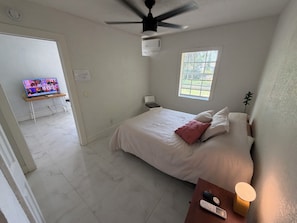 Een slaapkamer met een bed, een televisie op een standaard, een plafondventilator, een raam en een nachtkastje met een lamp.