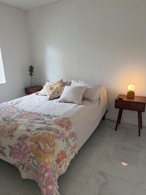 Een net opgemaakt bed met een bloemenkleed, een nachtkastje met een lamp en een kleine plant erop.