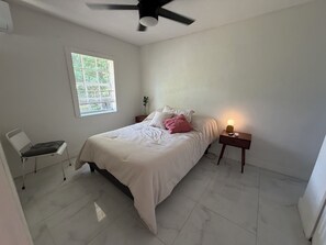 Een slaapkamer met een bed, nachtkastje, stoel, raam en plafondventilator.
