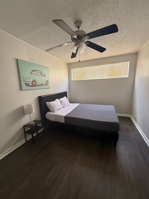 Een slaapkamer met een bed, een plafondventilator, een raam met zonwering, een nachtkastje met een lamp en een muur met een afbeelding van een auto.