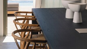 Een moderne kamer met een houten eettafel, rieten stoelen en uitzicht op een kustlandschap door een groot raam.