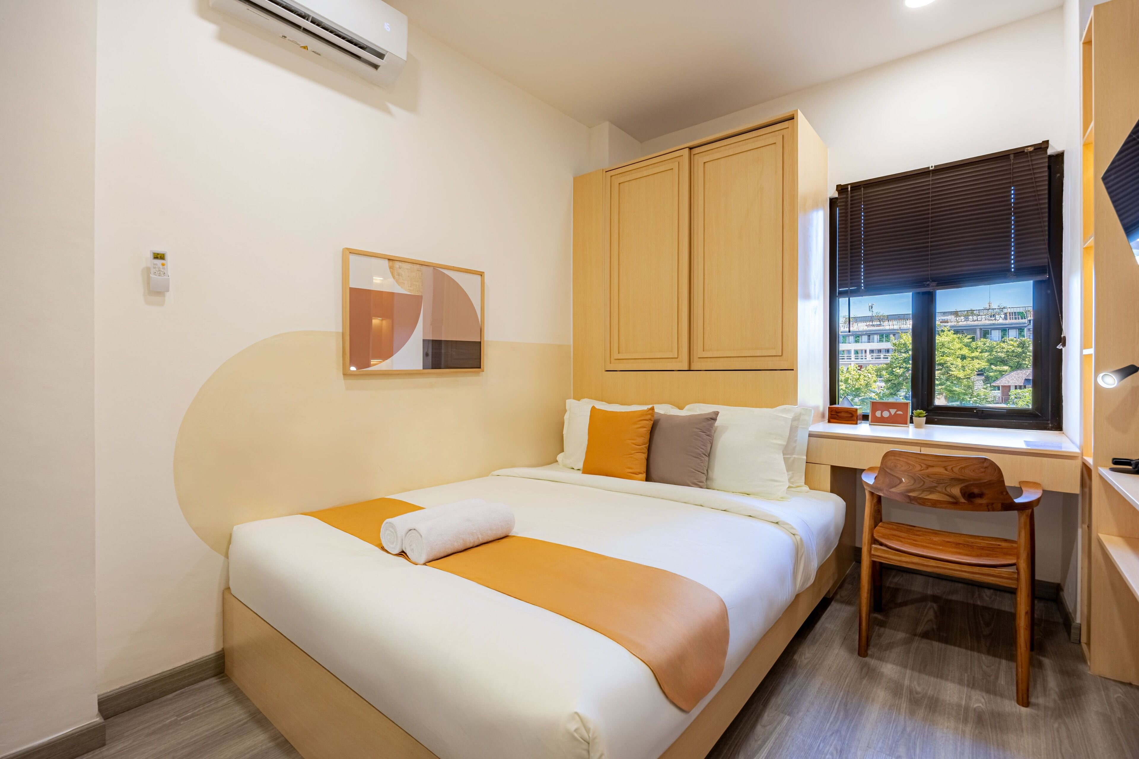A hotel room with a bed, a desk, a chair, a window with blinds, and a view of a building outside.