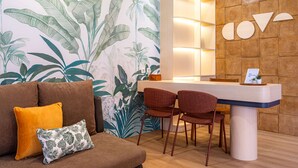 A reception desk with a wooden panel wall, a large plant mural, and a brown sofa with cushions. - Cove Meraya (Tibubeneng)