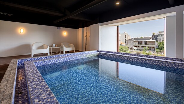 A modern indoor swimming pool with a tiled edge, a white chair, and a view of buildings outside. - Cove Meraya (Tibubeneng)