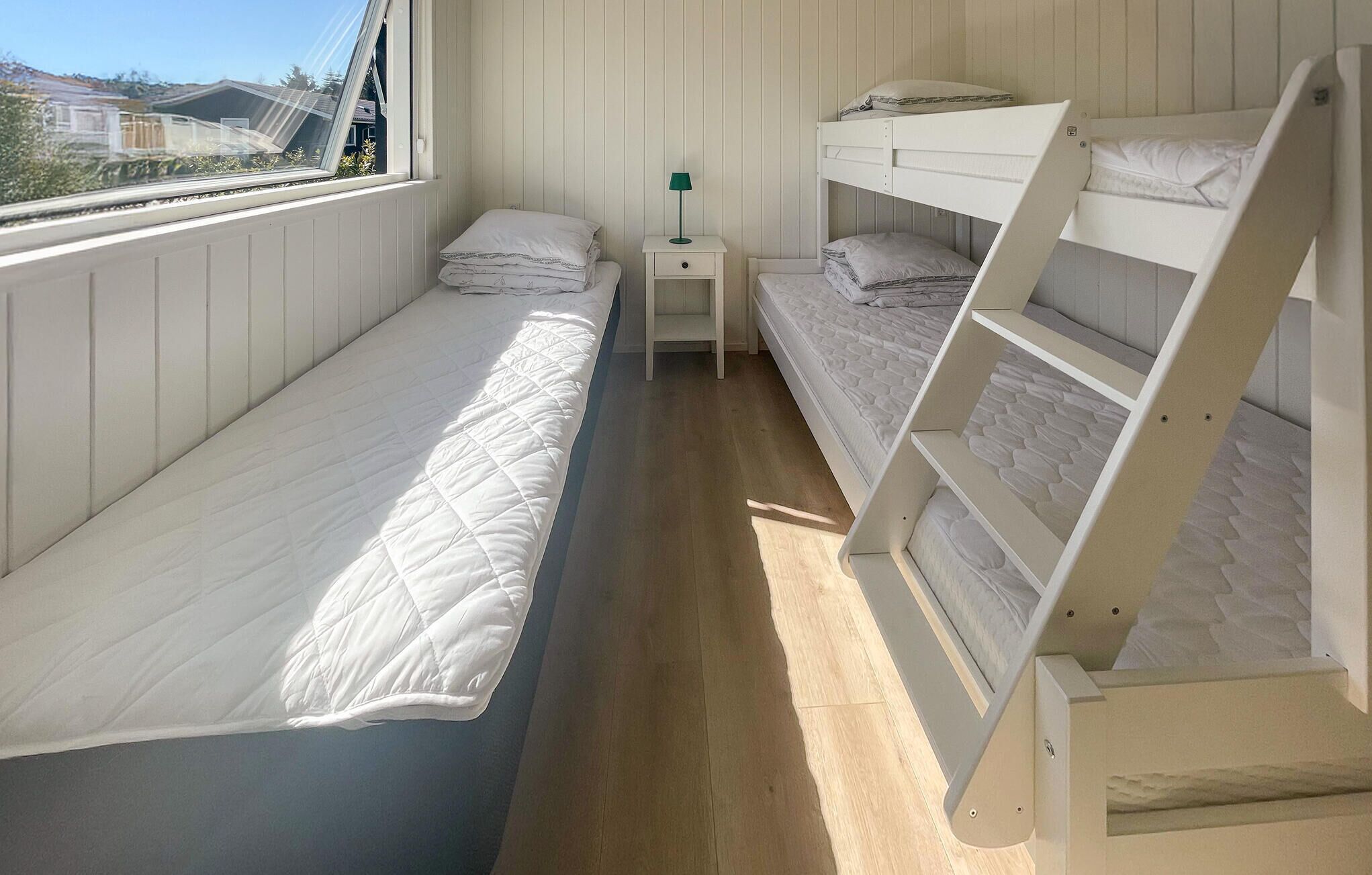 A room with three bunk beds, a small bedside table with a lamp, and a window with a view of houses.