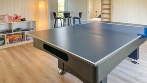 A ping pong table in a room with wooden flooring, a ladder, and a small table with chairs.