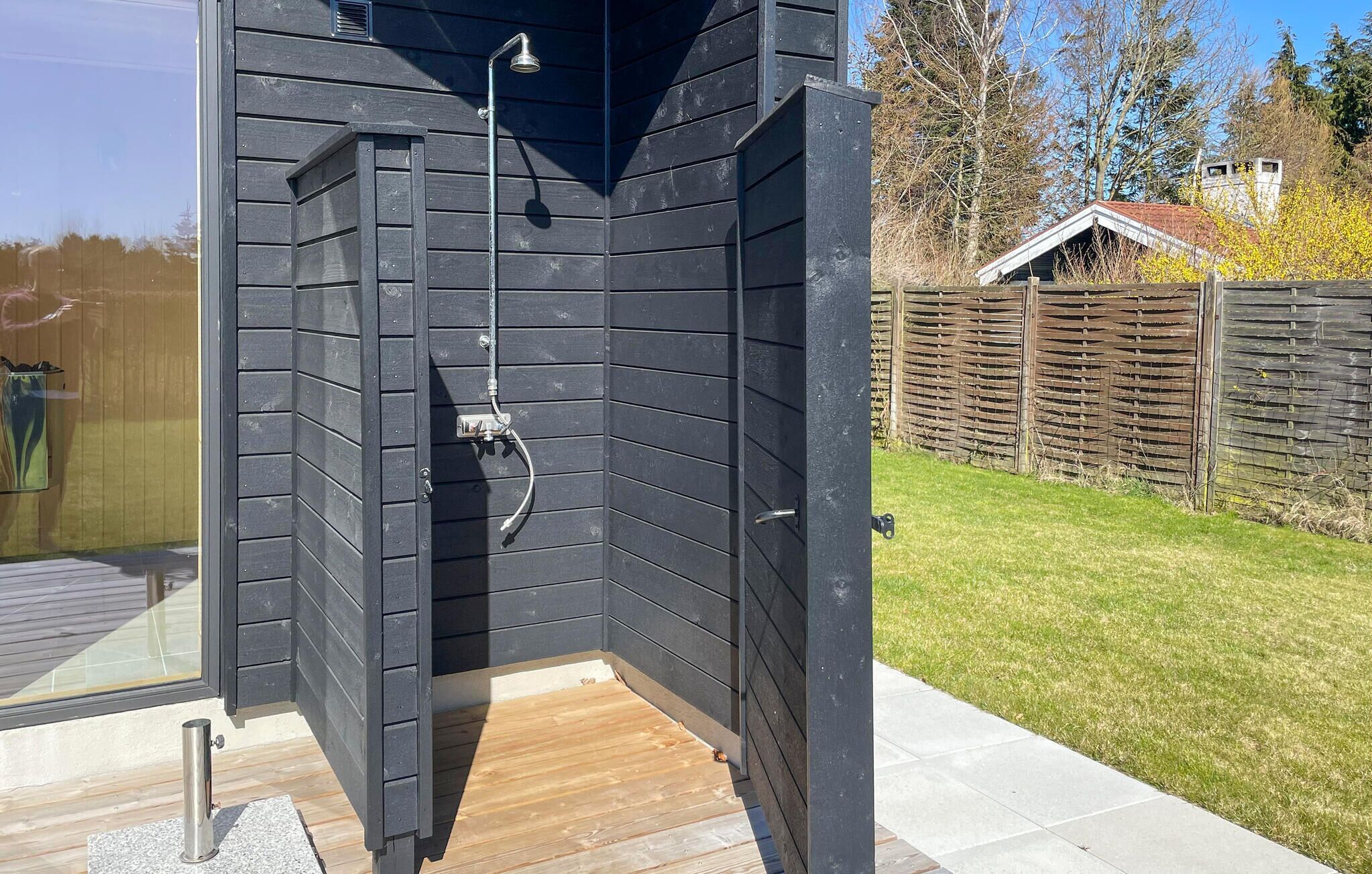 Een zwarte houten buitendouche met een houten terras, een metalen douchekop en een tuinslang.