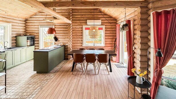 A log cabin interior with a dining area, kitchen, and a large window with red curtains.