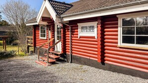 A red log cabin with a wooden staircase leading to a door, a window, and a gravel driveway.