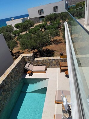 Uma área de piscina moderna com espreguiçadeiras, parede de pedra e vista para o mar e paisagem ao redor.