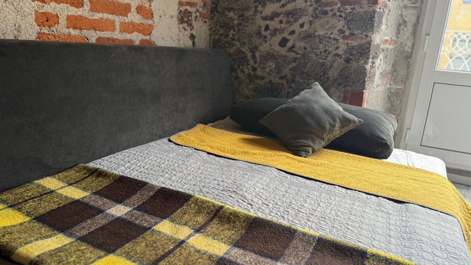 Una cama con cabecera gris, manta amarilla, almohada gris y manta a cuadros.