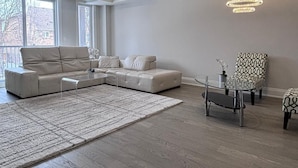 Ein modernes Wohnzimmer mit einem Ecksofa, einem gemusterten Sessel, einem Glastisch und einem großen Fenster.
