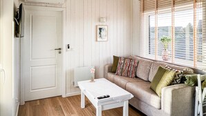 Ein Zimmer mit einer weißen Tür, einem weißen Tisch mit Fernbedienung, einem beigefarbenen Sofa mit gestreiften Kissen, einem gerahmten Bild an der Wand und einem Fenster mit Jalousien.