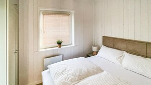 Ein Schlafzimmer mit einem Bett, einem Fenster mit Jalousien, einem Nachttisch mit Lampe und einer kleinen Pflanze auf der Fensterbank.