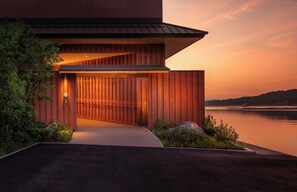 A modern building with a sloped roof and wooden paneling, situated by a body of water with a sunset in the background. - Hoshino Resorts KAI Miyajima (Hatsukaichi)
