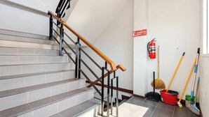 Una escalera con barandilla de madera, peldaños de baldosas grises, un extintor y utensilios de limpieza en el suelo.