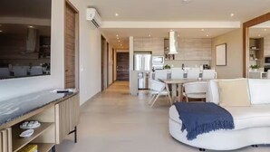 Uma sala de estar moderna com um sofá branco, uma mesa de centro e uma televisão. É possível ver uma cozinha ao fundo, com uma área de jantar.