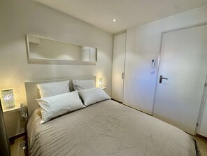 A bedroom with a bed, a bedside table, a nightstand, a wall-mounted mirror, and a door. - IN THE HEART OF THE CITY CENTER, ONE BEDROOM APARTMENT FOR 4 PEOPLE (STE MAXIME)