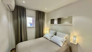 A hotel room with a bed, curtains, a window, an air conditioning unit, a wall-mounted mirror, and bedside lamps. - IN THE HEART OF THE CITY CENTER, ONE BEDROOM APARTMENT FOR 4 PEOPLE (STE MAXIME)