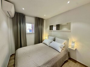 A hotel room with a bed, curtains, a window, an air conditioning unit, a wall-mounted mirror, and bedside lamps. - IN THE HEART OF THE CITY CENTER, ONE BEDROOM APARTMENT FOR 4 PEOPLE (STE MAXIME)