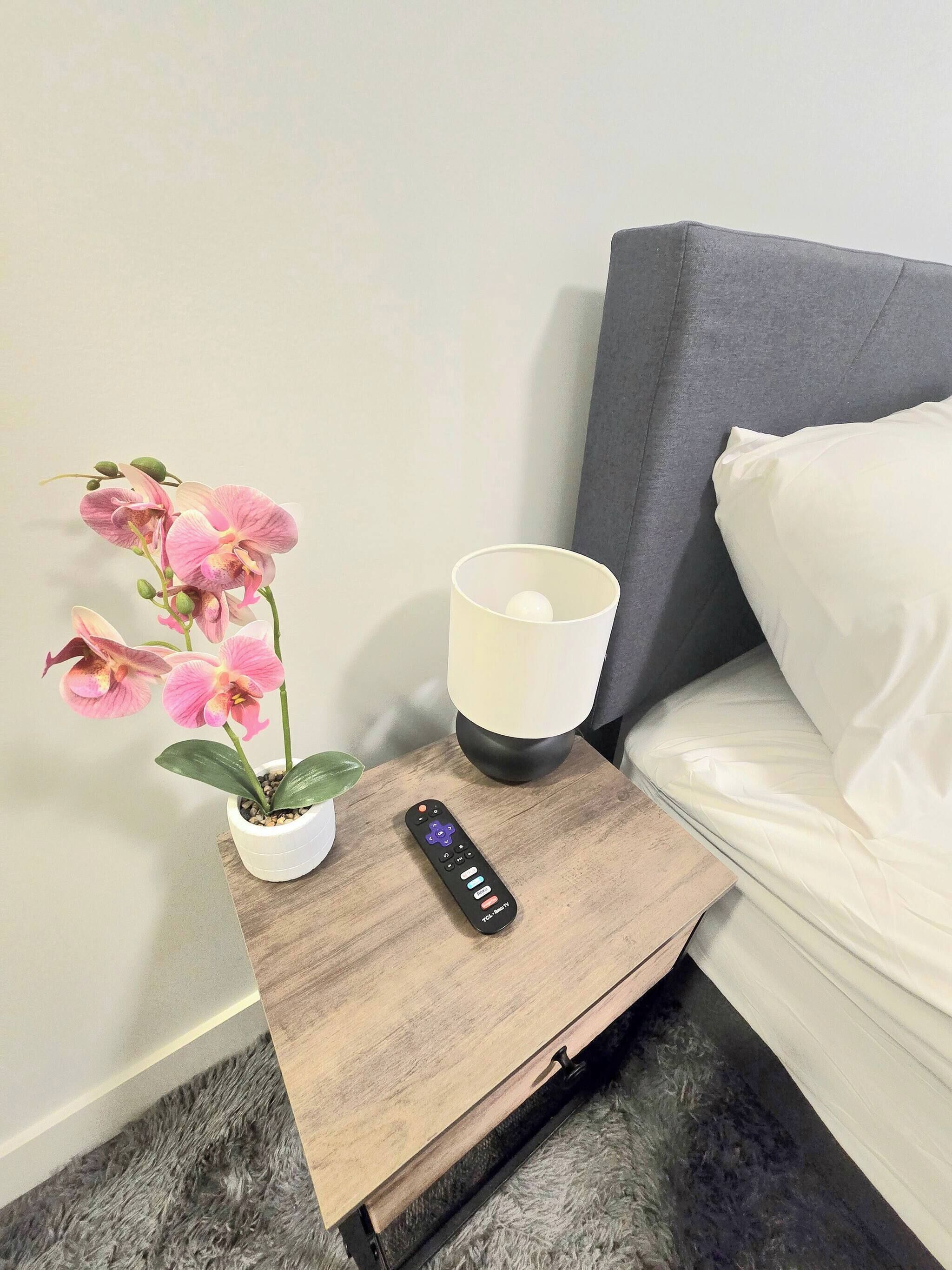 A bed with a grey headboard, a white pillow, a small wooden nightstand with a potted orchid, a remote control, and a white lamp.