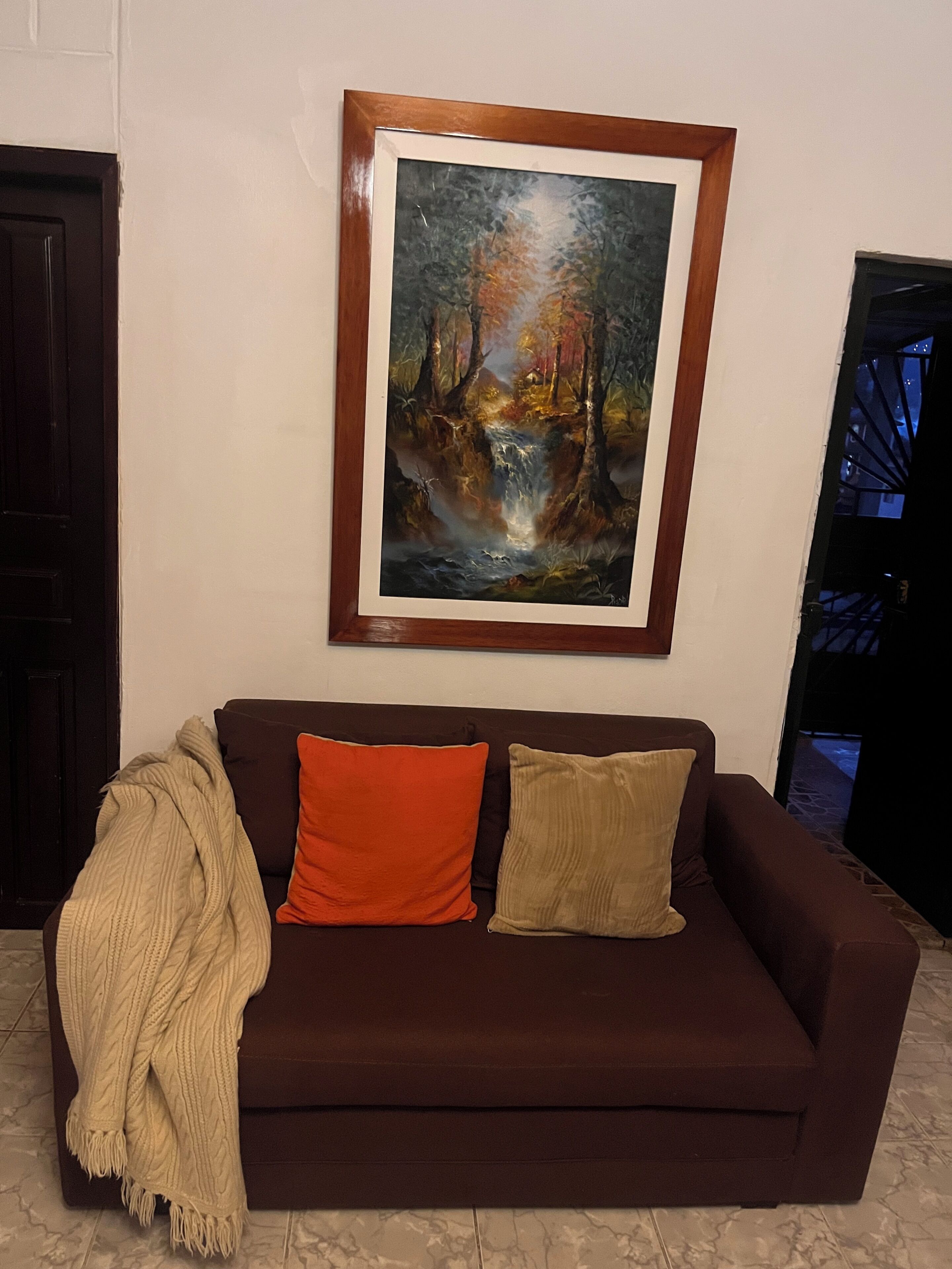 A brown sofa with two cushions, a blanket, and a framed painting of a forest scene.