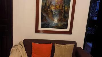 A brown sofa with two cushions, a blanket, and a framed painting of a forest scene.