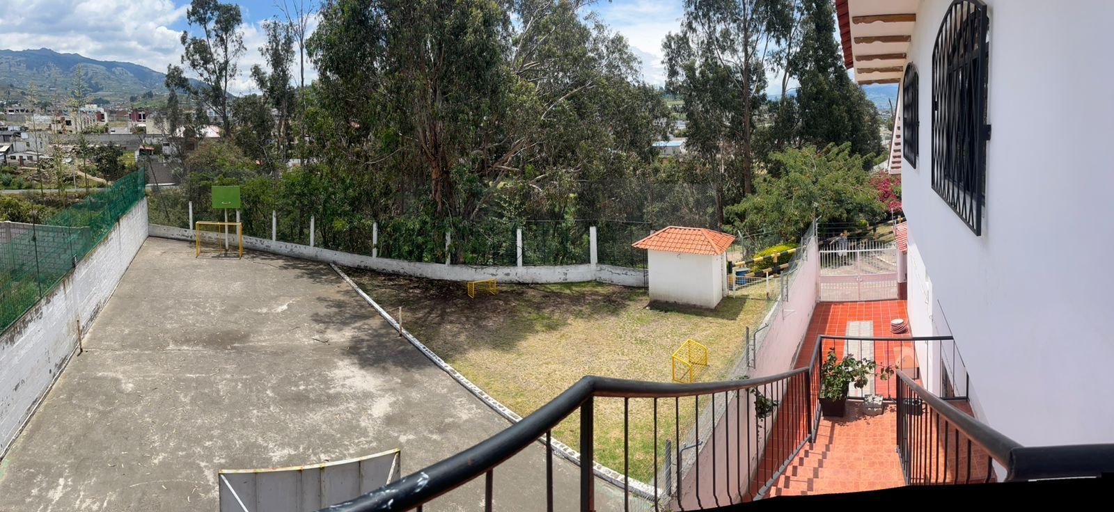 A residential area with a view of a fenced yard, a small building with a red roof, and a balcony with a railing.