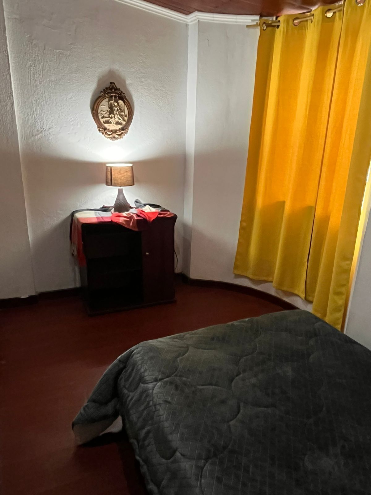 Ein Zimmer mit einem Bett, einem Schreibtisch mit Lampe, einem kleinen Schrank und einem gelben Vorhang.