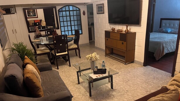 A living room with a sofa, a coffee table, a dining area with chairs, a TV mounted on the wall, and a bed in the adjacent room.