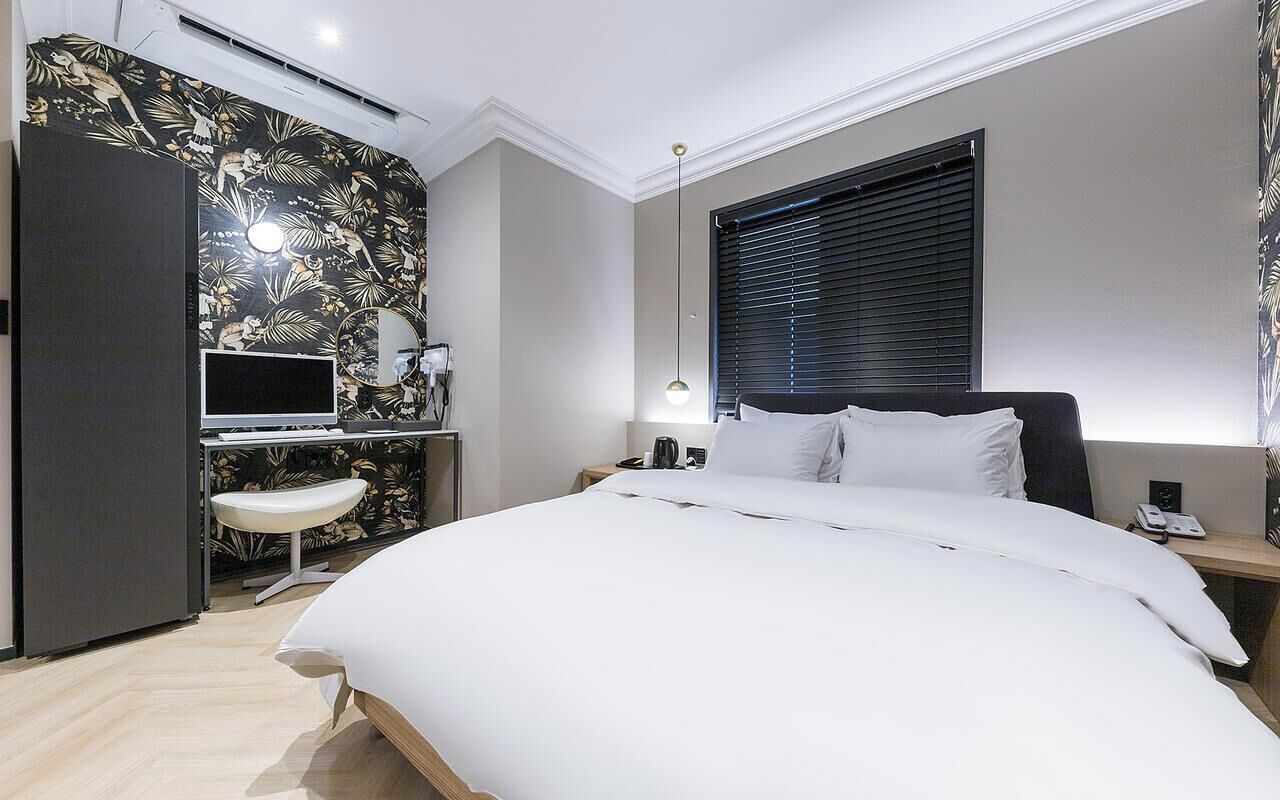 Hotellihuone, jossa on sänky, työpöytä tietokoneella, tuoli, ikkuna, jossa on sälekaihtimet, ja seinälle asennettu ilmastointilaite.