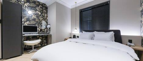 Hotellihuone, jossa on sänky, työpöytä tietokoneella, tuoli, ikkuna, jossa on sälekaihtimet, ja seinälle asennettu ilmastointilaite.