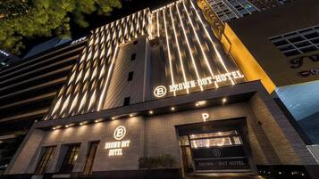 Un hotel moderno con un llamativo letrero 'BROWN-DOT HOTEL', una entrada de estacionamiento y una fachada con detalles de iluminación vertical.