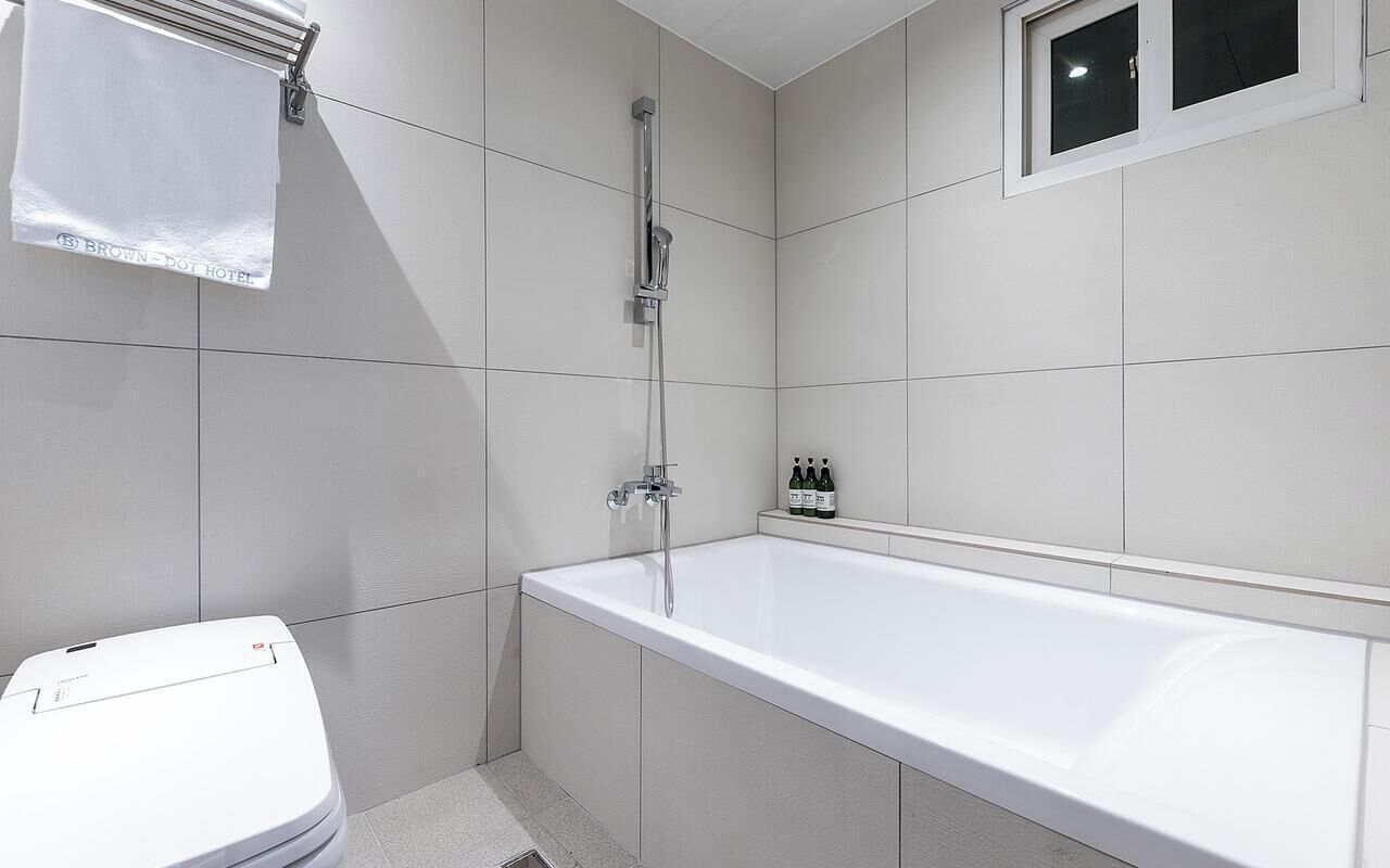 Une salle de bain moderne avec une baignoire blanche, une douche et des toilettes. Une serviette est accrochée à un porte-serviettes et une petite fenêtre se trouve au-dessus de la baignoire.