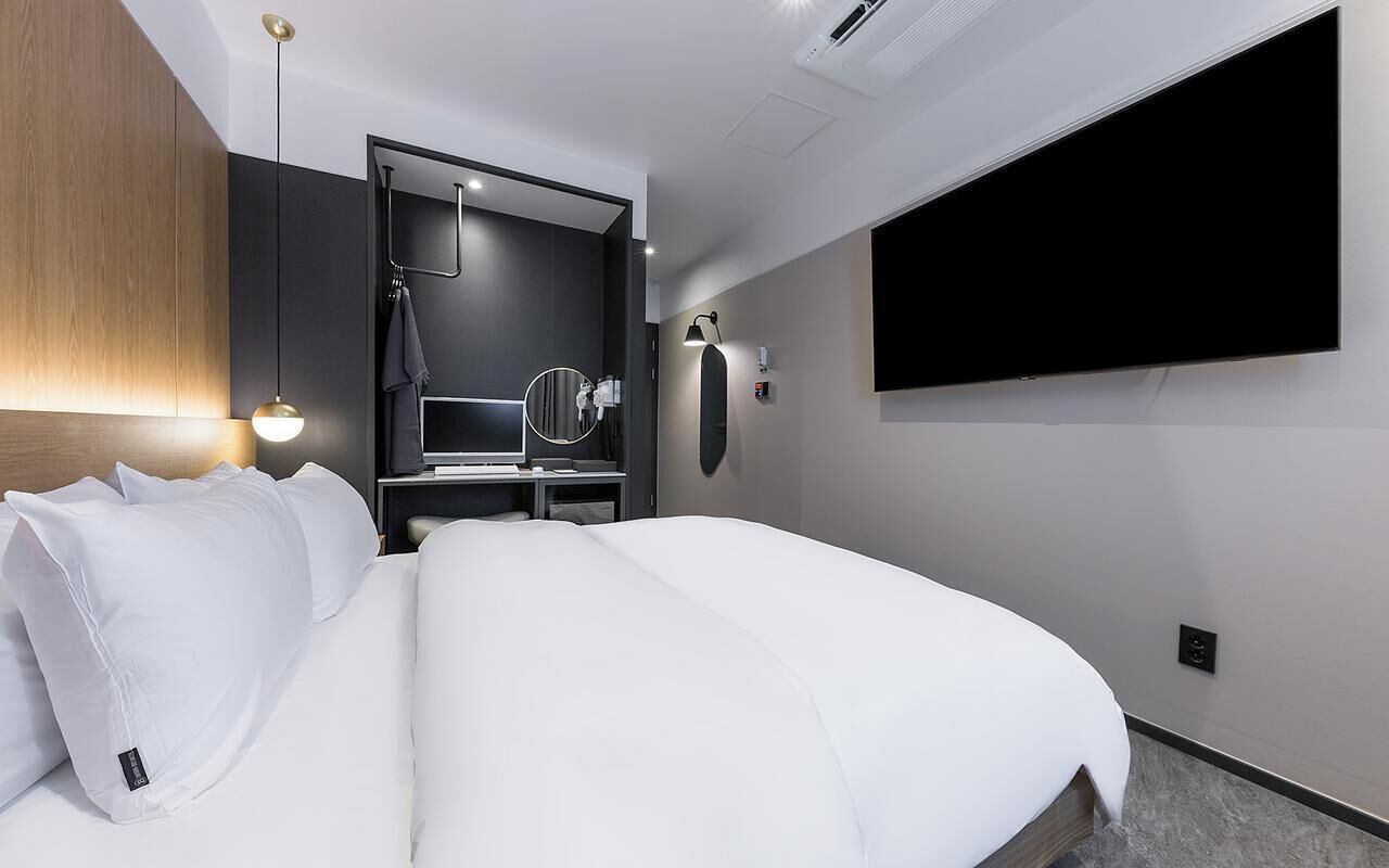 A modern hotel room with a large bed, a TV mounted on the wall, a mirror, and a small table with a lamp.
