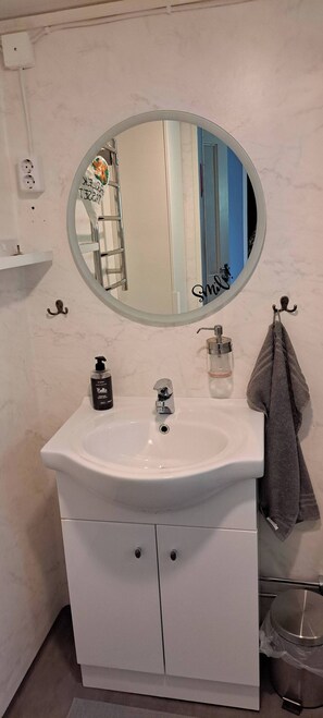 A bathroom with a white sink, a round mirror, a towel rack, a soap dispenser, and a trash bin. - Welcome to Lillstugan (Jämshög)
