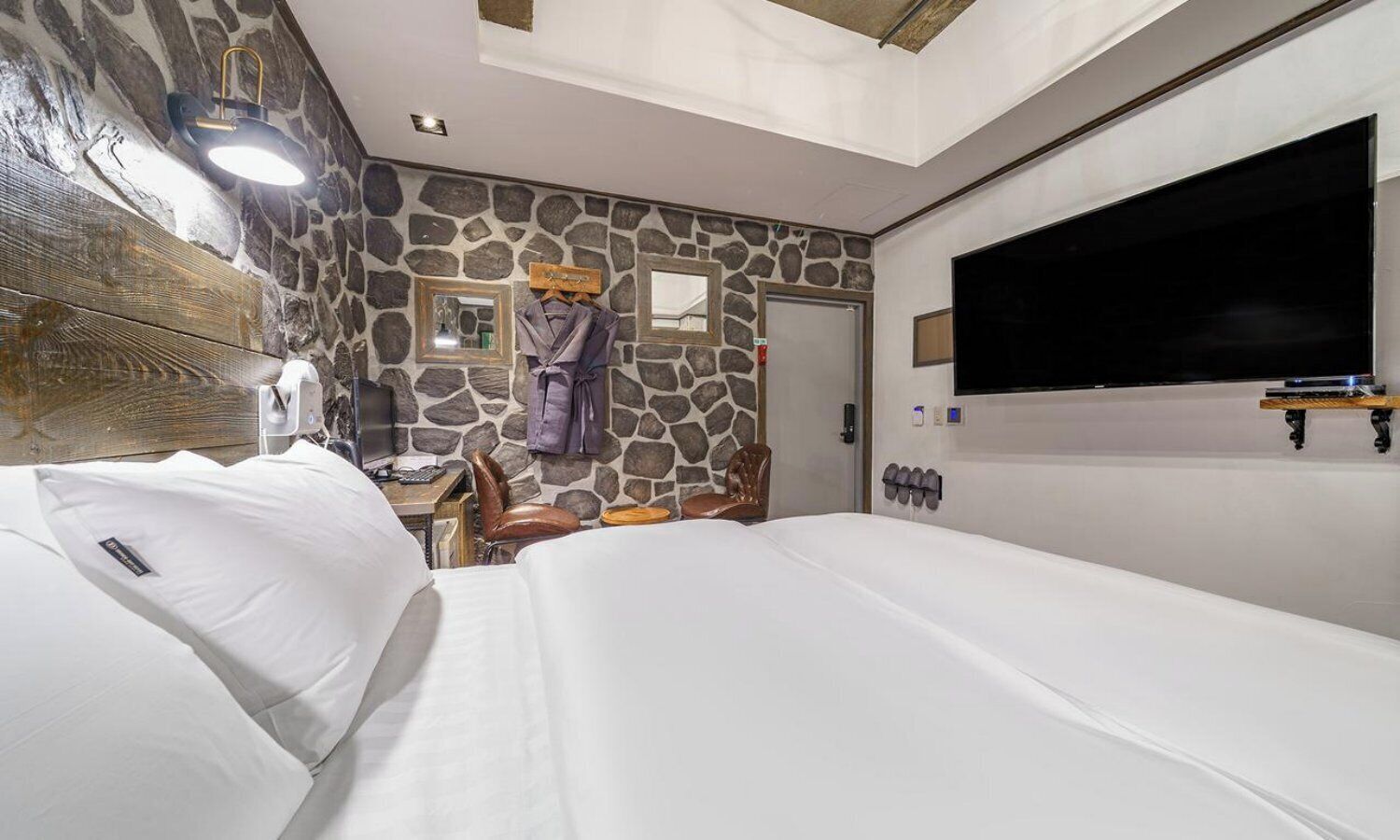 Habitación de hotel con pared de piedra, una cama, un escritorio con computadora, televisor y espejo.