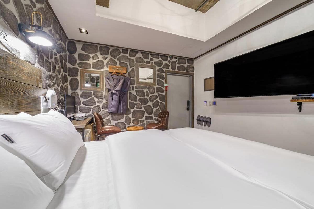 Habitación de hotel con pared de piedra, una cama, un escritorio con computadora, televisor y espejo.