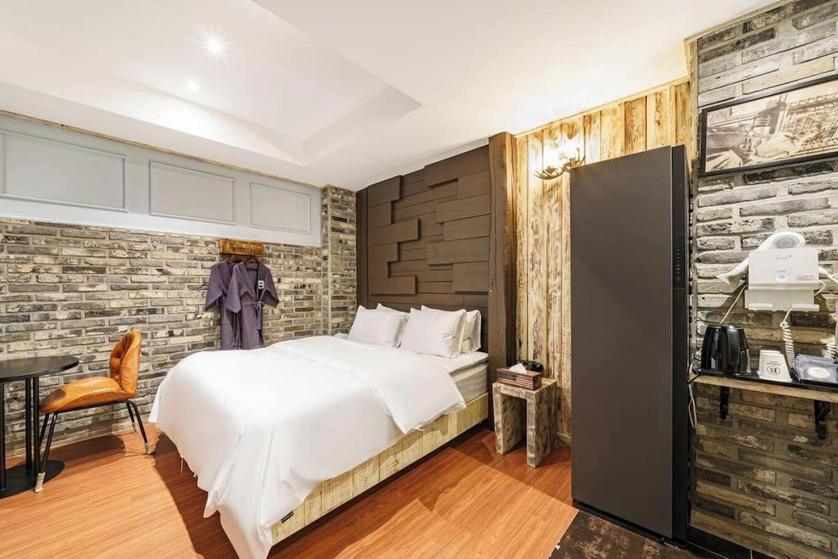 Habitación de hotel con pared de ladrillo, cama con ropa blanca, piso de madera, una pequeña mesa con una silla y un perchero con bata.
