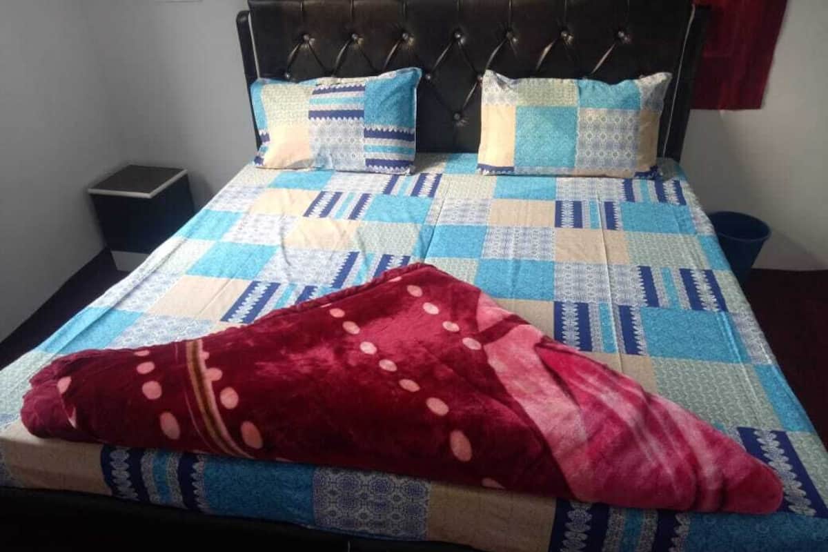 Uma cama com colcha e duas almofadas, uma mesinha de cabeceira e uma lixeira.