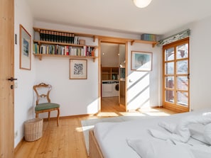 Un dormitorio con una cama, una silla, una estantería y vistas a una cocina a través de una puerta abierta.