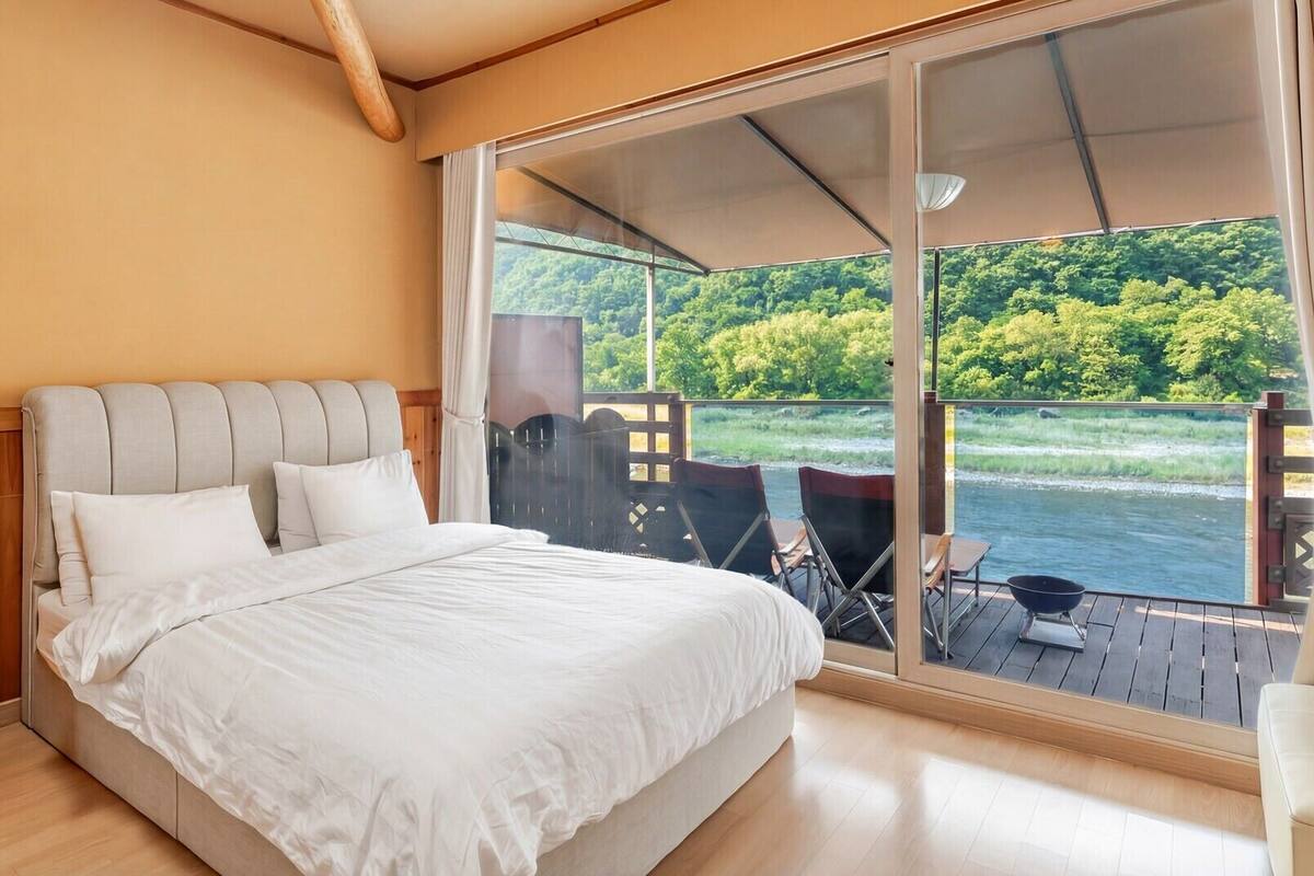 A bedroom with a large bed, a view of a river, and a deck with chairs and a grill.