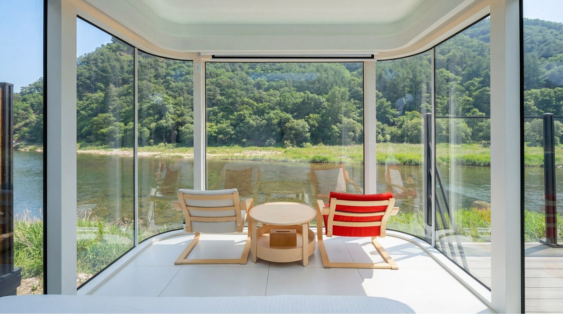 A glass-enclosed room with a table and chairs, overlooking a river and forest.