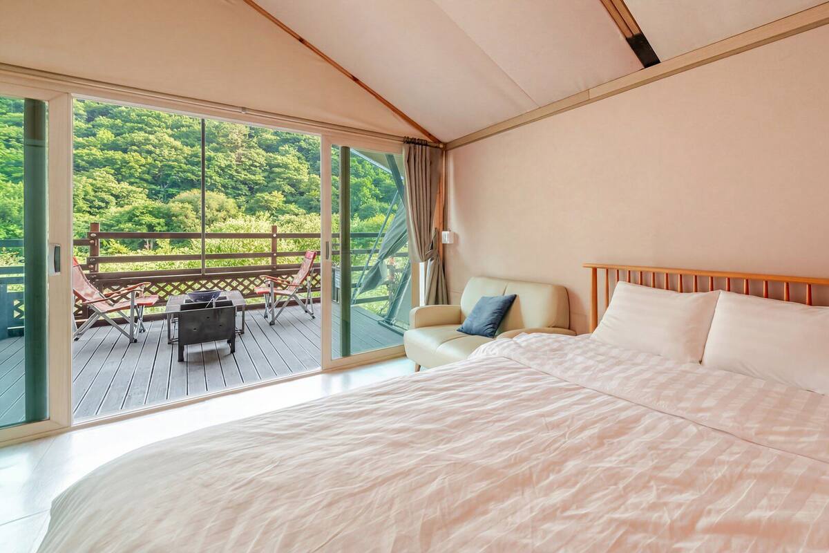 A bedroom with a large bed, a sofa, and a balcony with a view of greenery.