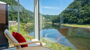 Une pièce avec vue sur une rivière et une forêt, une chaise avec un coussin et un plancher en bois.