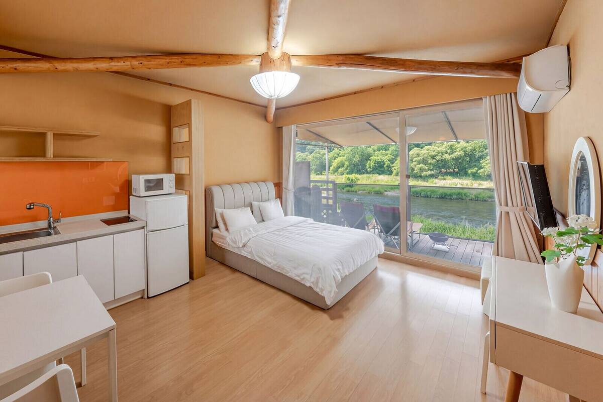 A compact room with a bed, a small kitchenette, a dining table, and a large window with a view of greenery.