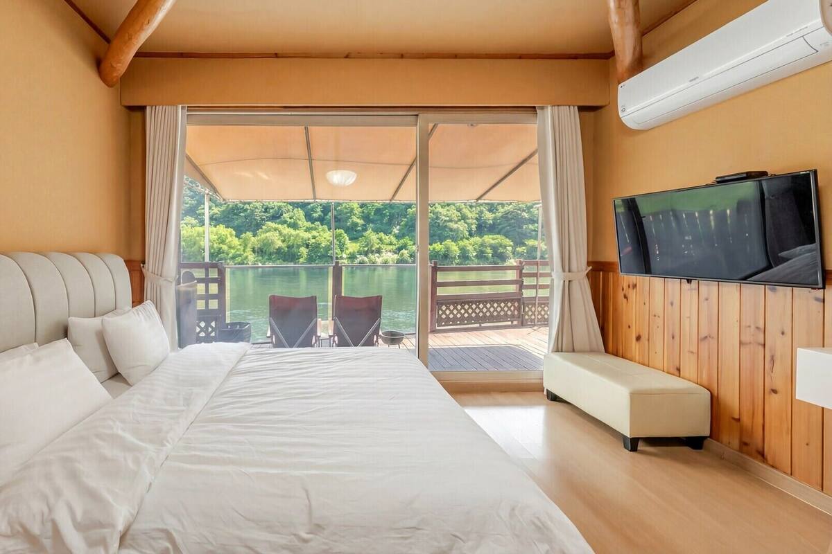 A bedroom with a bed, a TV, and a balcony with a view of greenery.