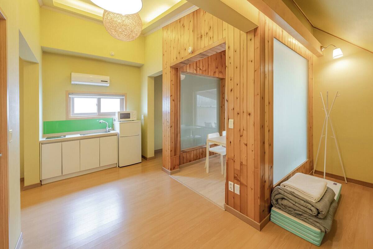 A room with a wooden interior, a kitchenette with a microwave, a sink, and cabinets, a glass door leading to another room, and a bed with a folded blanket.