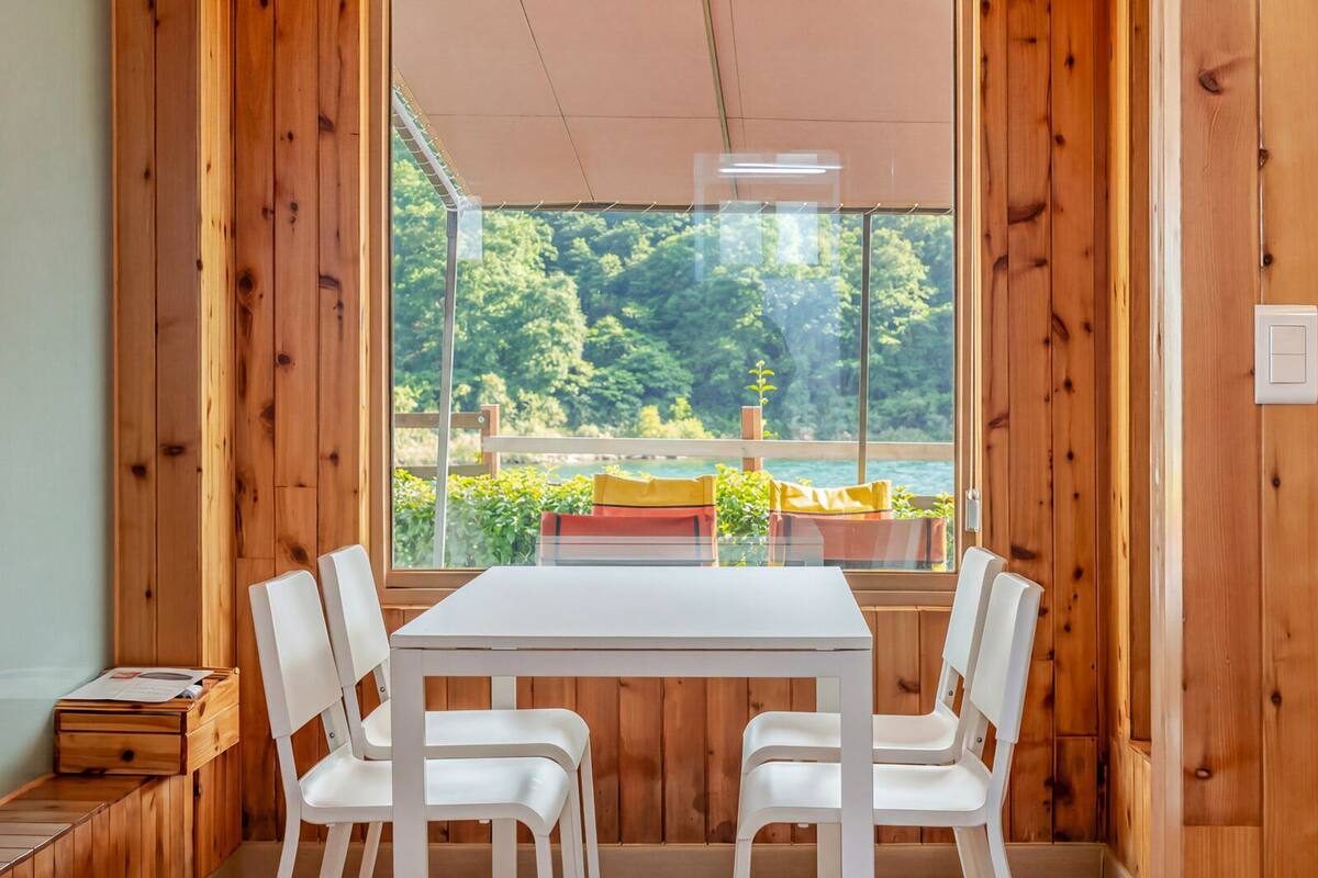 A wooden-paneled room with a white table and chairs, a window with a view of greenery, and a small wooden bench.