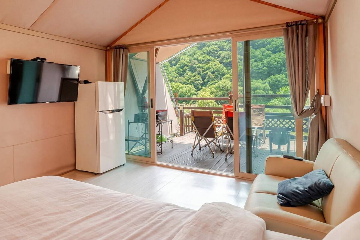 A bedroom with a bed, a TV, a sliding glass door to a balcony, and a view of greenery.